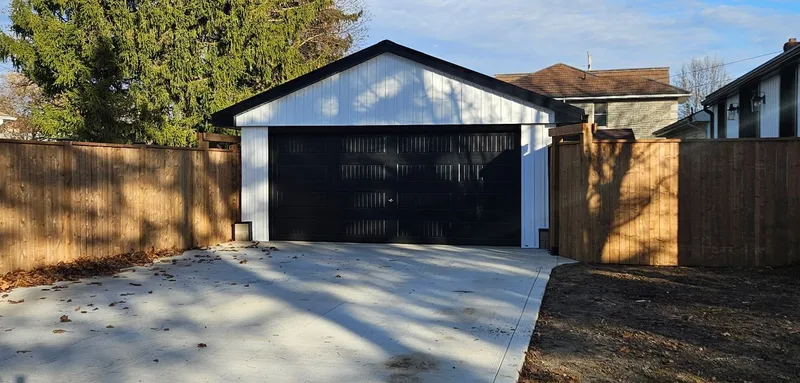 Front view of a white garage with a black overhead door, beside a driveway and wood fence